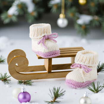 Handgestrickte Babyschuhe in Cremeweiß mit fliederfarbener Sohle und Schleife auf kleinem Holzschlitten im warmen Sonnenlicht