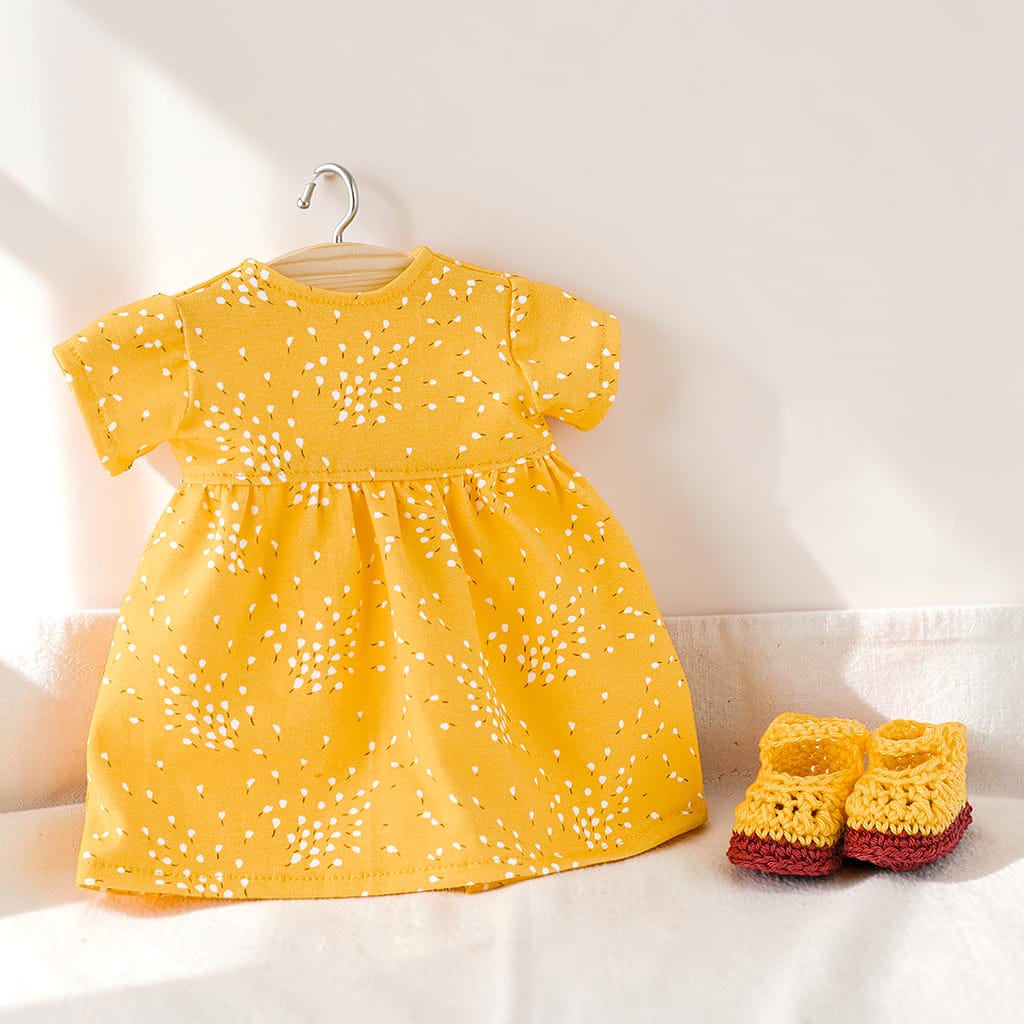 Gelbes Puppenkleid mit kurzen Ärmeln und weißem Blütenmuster auf einem Kleiderbügel, dazu gehäkelte Puppenschuhe