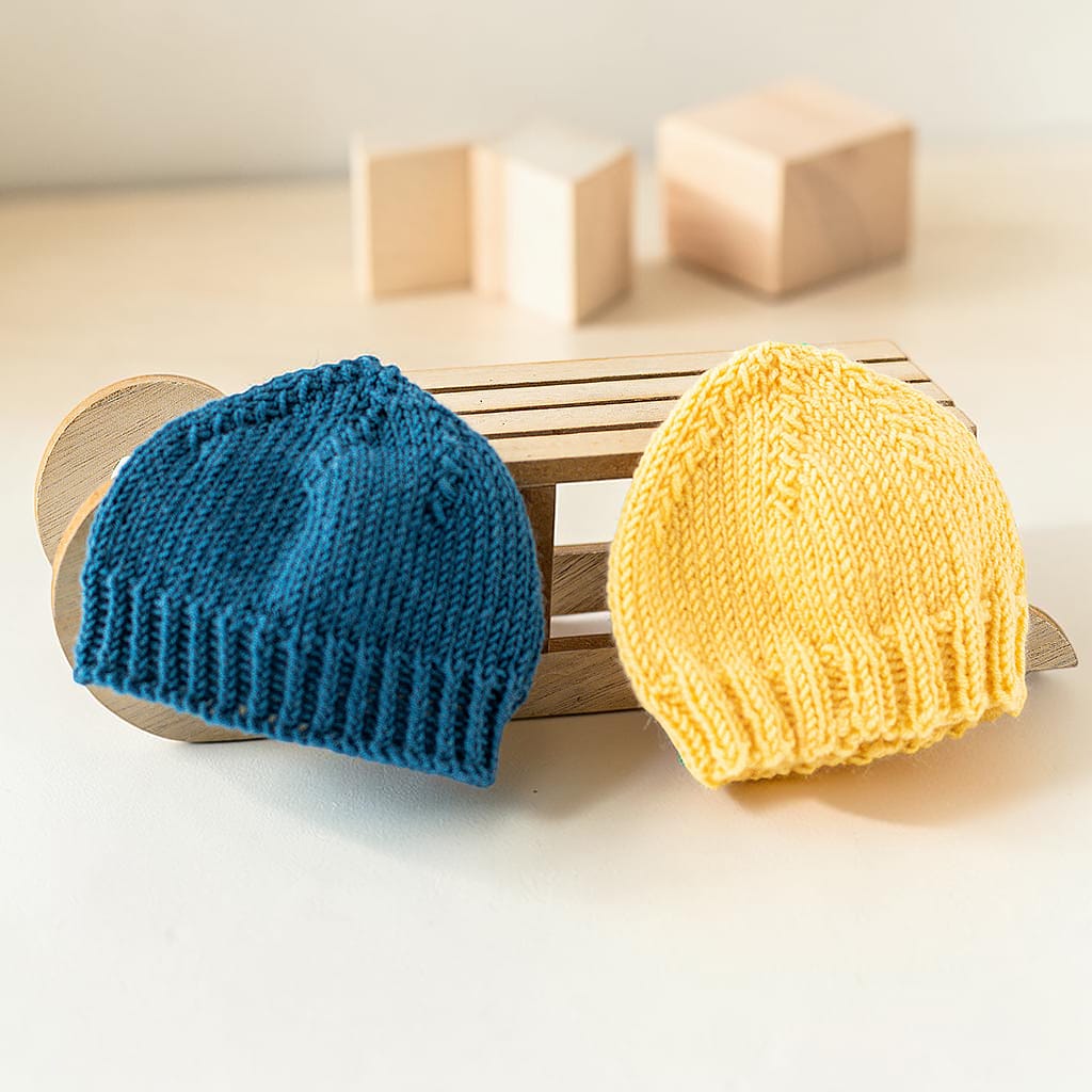 Handgestrickte Puppenmützen aus Wolle in Petrol und Senfgelb auf Holzschlitten fotografiert