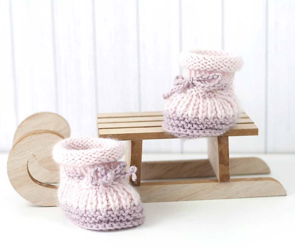 Gestrickte Puppenschuhe aus Merinowolle in Rosa und Grau neben kleinem Holzschlitten