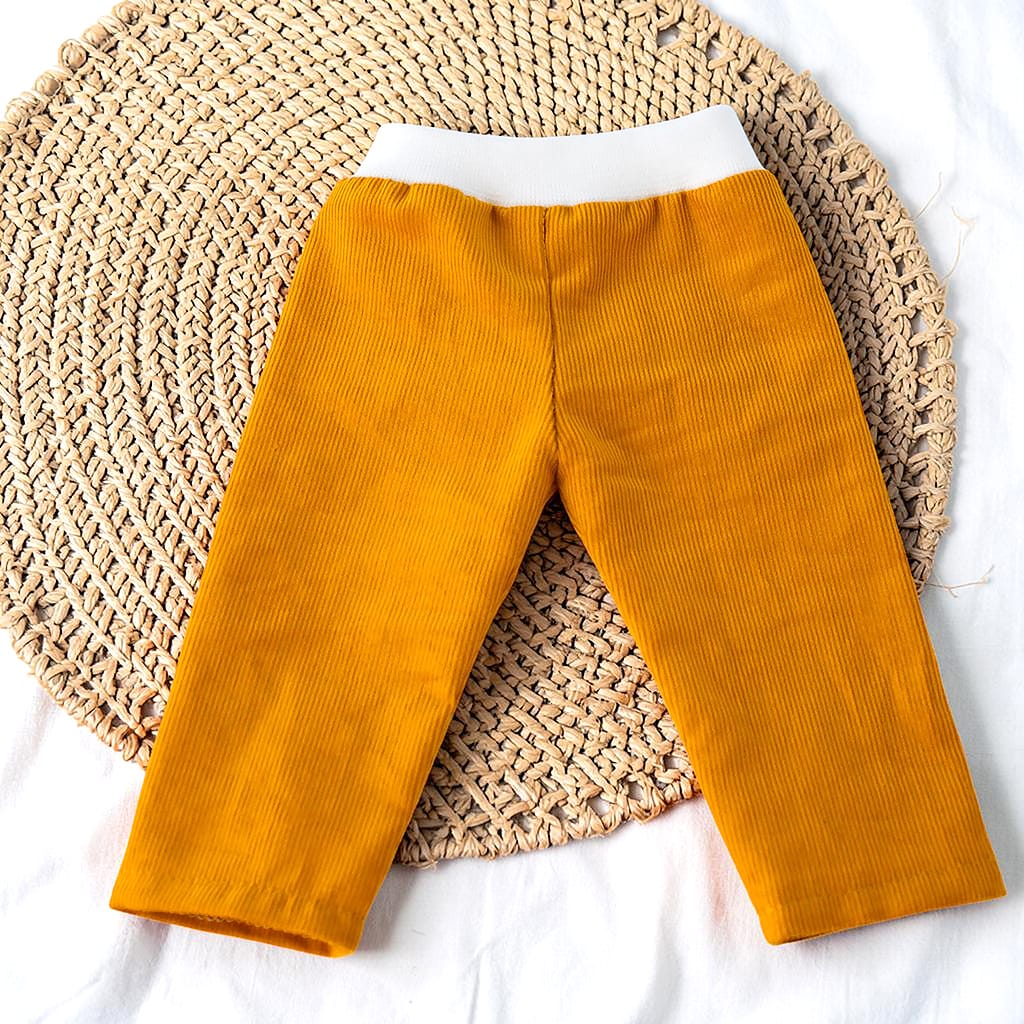 Senfgelbe Puppenhose aus Babycord mit weißem Bündchen, flach auf einer runden Flechtmatte arrangiert. Die feine Cordstruktur und die gerade Passform sind gut sichtbar.