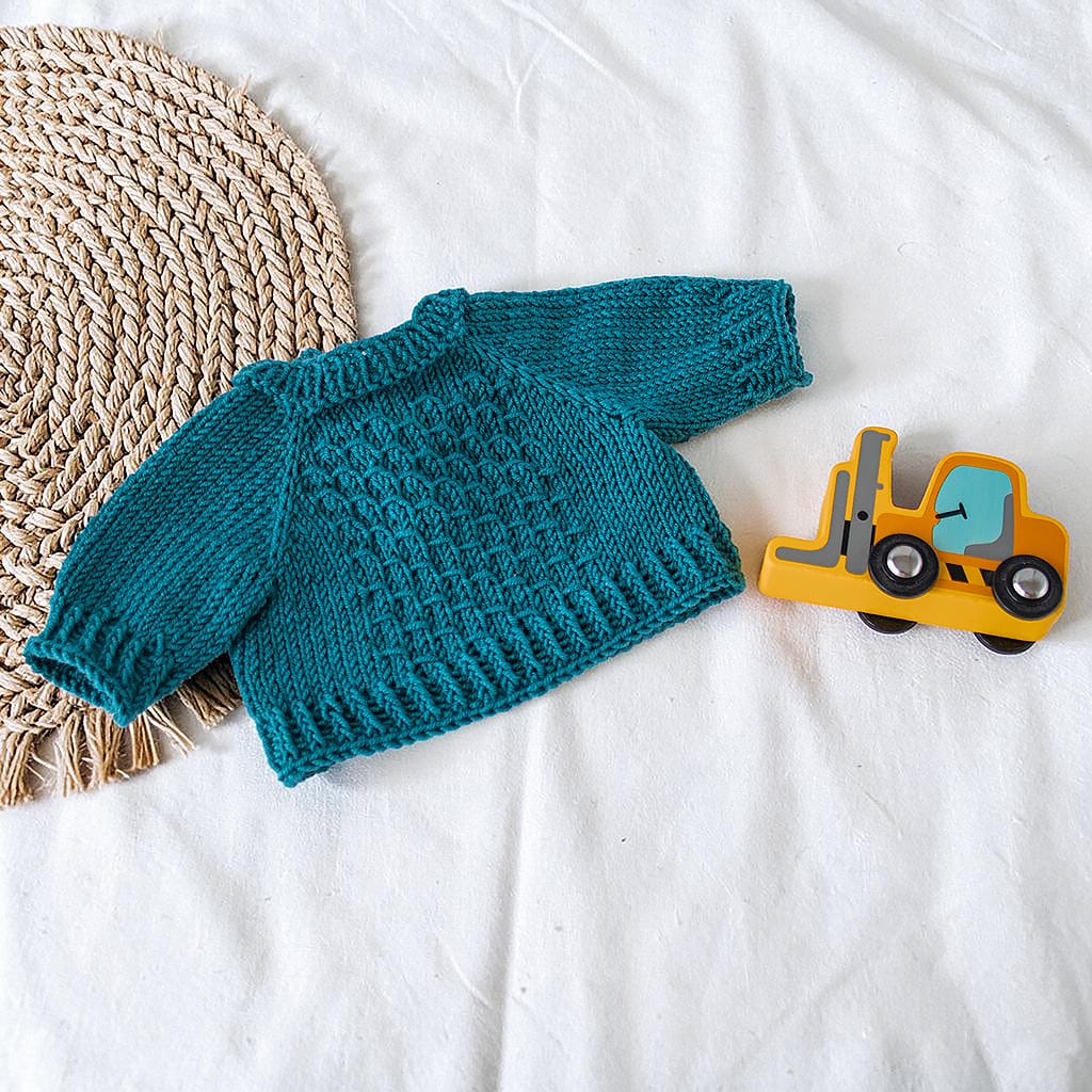 Handgestrickter Puppenpullover aus Merinowolle in Petrol für Waldorfpuppen, fotografiert auf hellem Stoff mit Flechtmatte und kleinem Holzbagger.