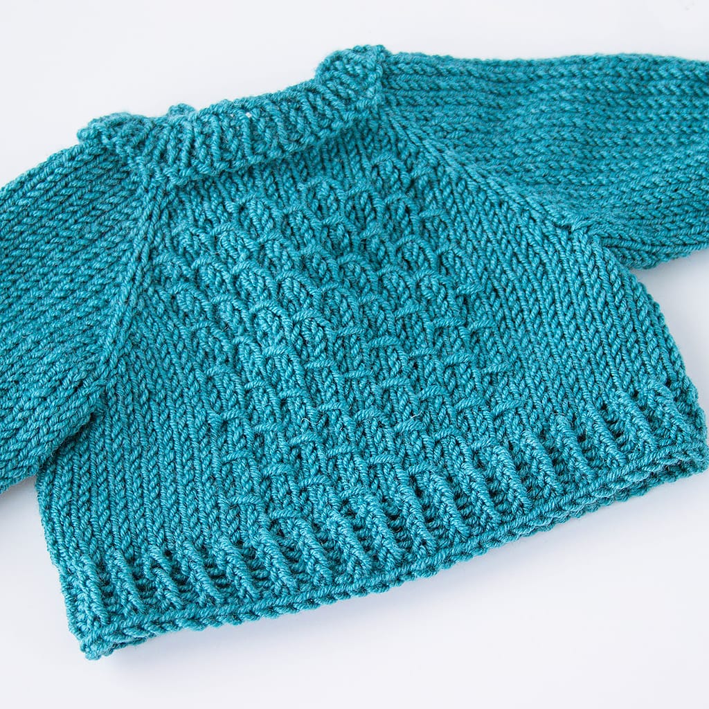 Detailaufnahme des handgestrickten Strukturmusters eines petrolfarbenen Puppenpullovers aus Merinowolle.