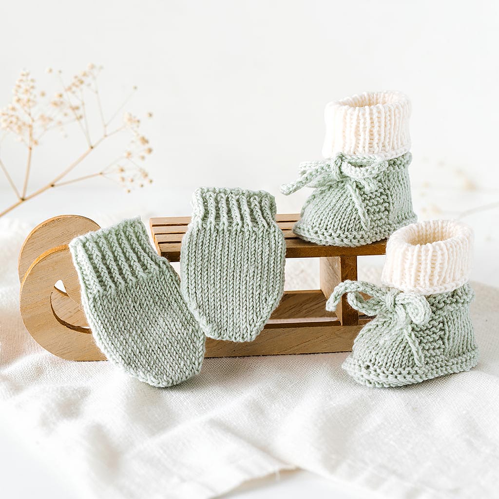 Handgestricktes Babyset aus salbeigrünen Babyschuhen und Fäustlingen aus Merinowolle auf einem Holzschlitten dekoriert.