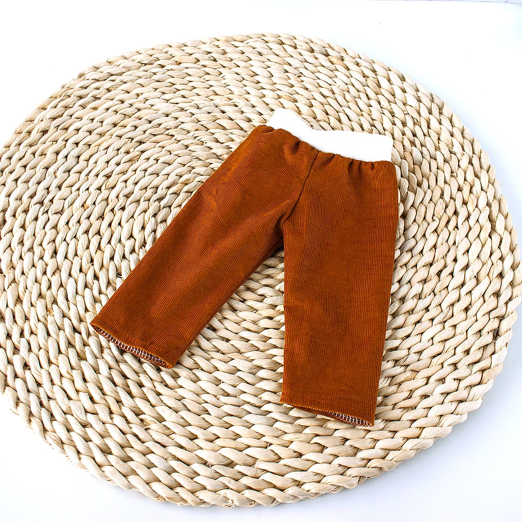 Rostbraune Puppenhose aus feinem Cord mit weichem weißem Bündchen für Puppen von 36–38 cm auf geflochtener Naturunterlage.