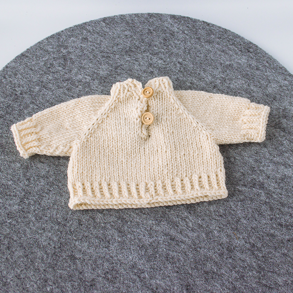 Rückseite eines handgestrickten Puppenpullovers aus naturfarbener Merinowolle mit Rollkragen und Knopfleiste aus zwei Holzknöpfen für Puppen von 36–40 cm.