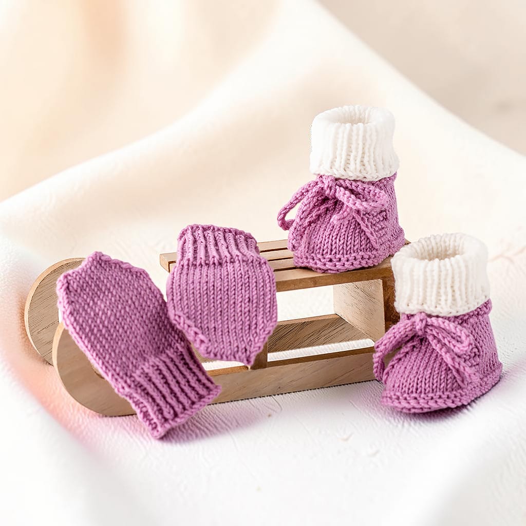 Gestricktes Babyset in Altrosa und Creme mit Babyschuhen und passenden Handschuhen auf einem Holzschlitten