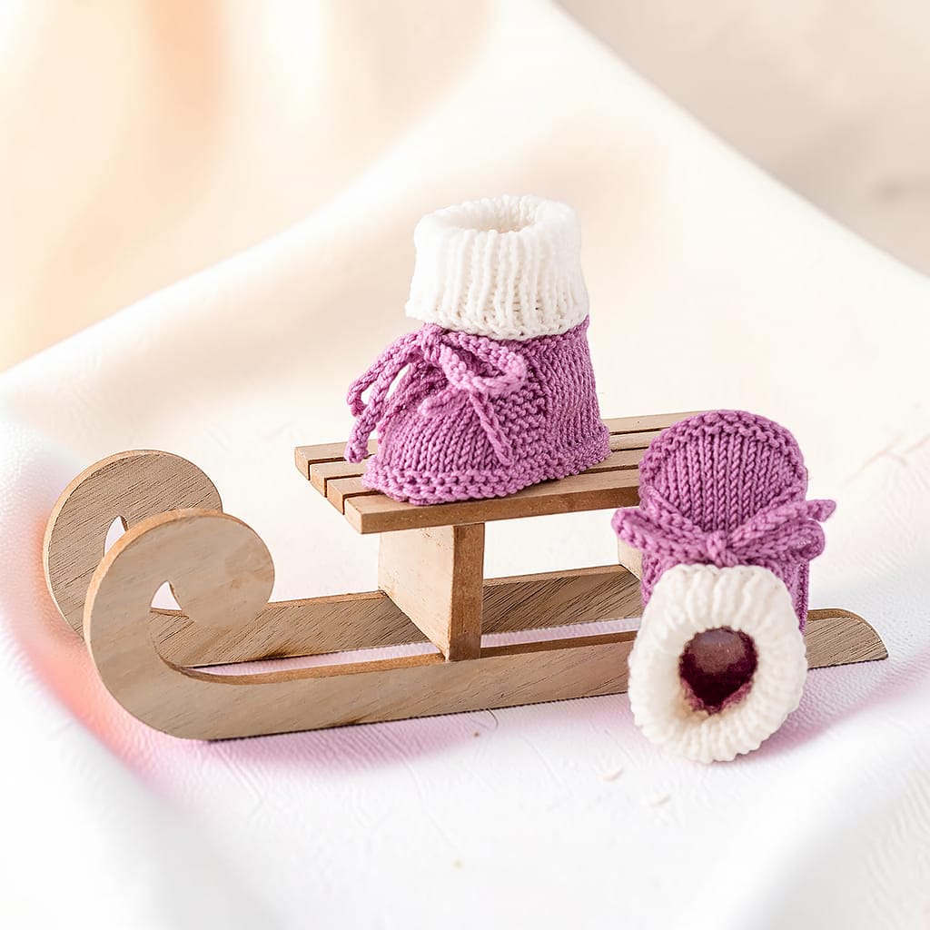 Gestrickter Babyschuh in Altrosa und Weiß neben einer aufgestellten Sohle, Ansicht von vorne und hinten