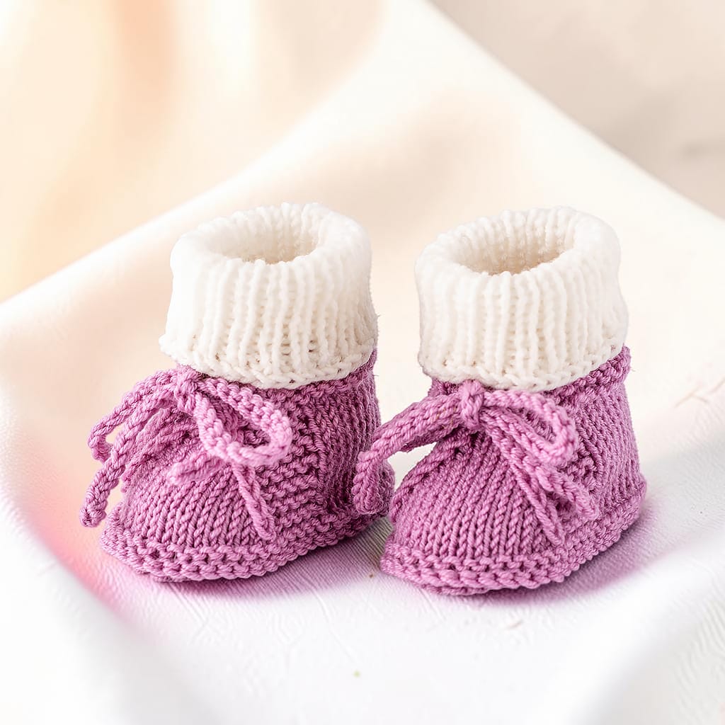 Paar gestrickte Babyschuhe in Altrosa mit weißem Umschlag und dekorativen Bindebändern