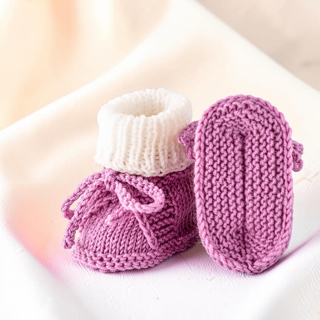 Gestrickte Babyschuhe in Altrosa und Weiß auf einem Holzschaukelpferd, eine Sohle zeigt die Verarbeitung von unten