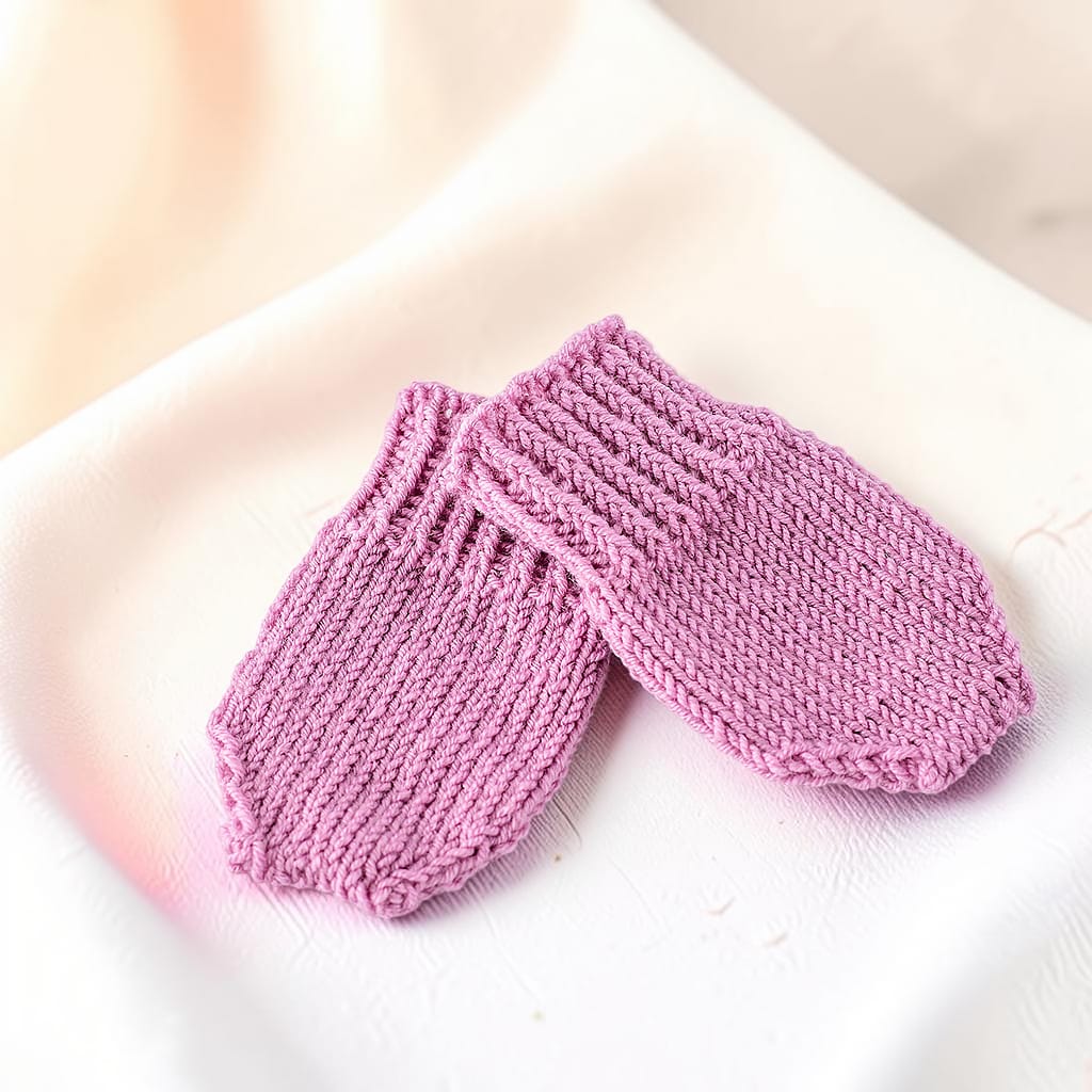 Zwei gestrickte Babyhandschuhe in Altrosa, übereinandergelegt und aus seitlicher Perspektive fotografiert