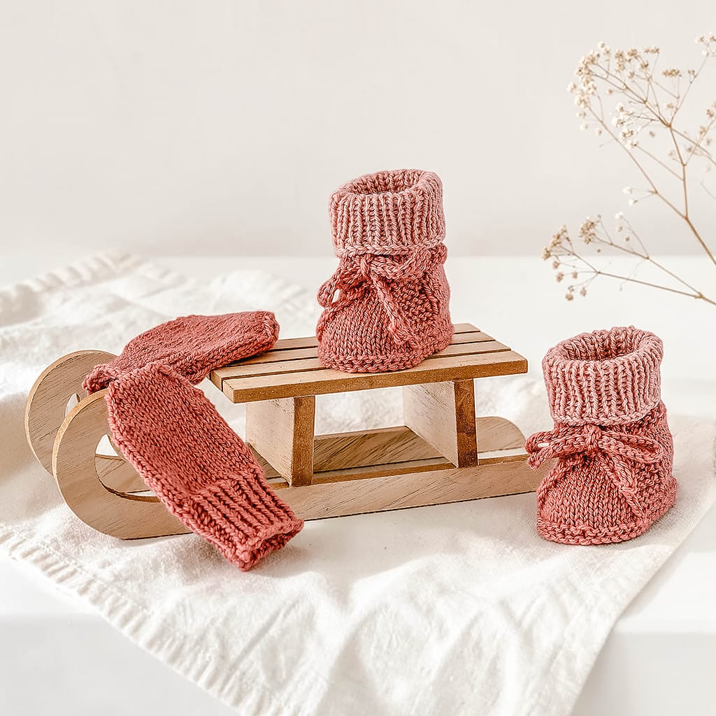 Handgestrickte Babyschuhe und Fäustlinge aus 80 % superfeiner Merinowolle und 20 % Kaschmir in Terrakotta-Rosé auf einem kleinen Holzschlitten dekoriert.