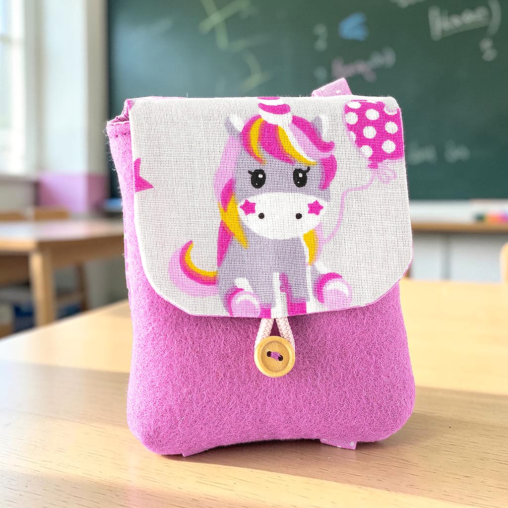 Puppenrucksack für Waldorfpuppen aus rosa Wollfilz mit Baumwollstoff in hellgrau mit einem sitzenden Einhorn. Der Rucksack hat einen Verschluss mit Kordel und Holzknopf.