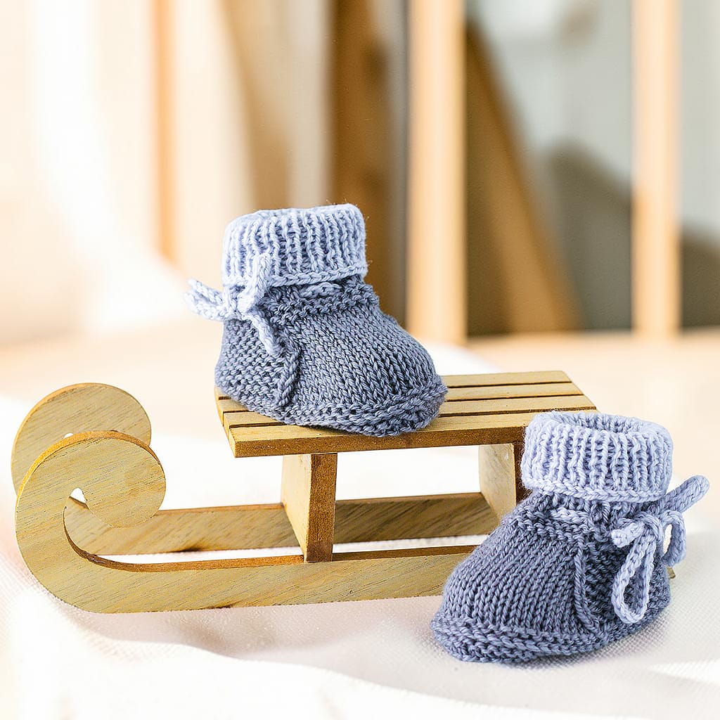 Graue gestrickte Babyschuhe und Fäustlinge aus Merinowolle und Kaschmir auf einem Holzschlitten